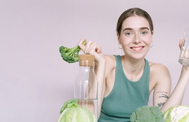Woman in tank top smiling with broccoli, cabbage, and glass of water on table.