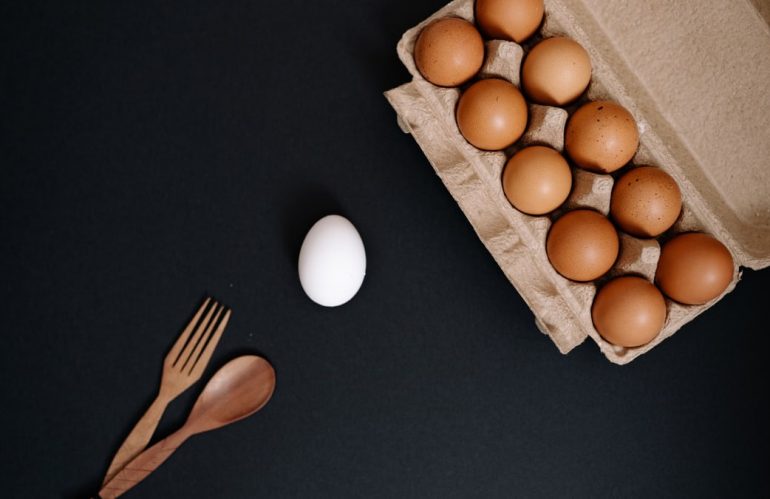 Brown eggs in carton with single white egg on dark background. Minimalist food concept.