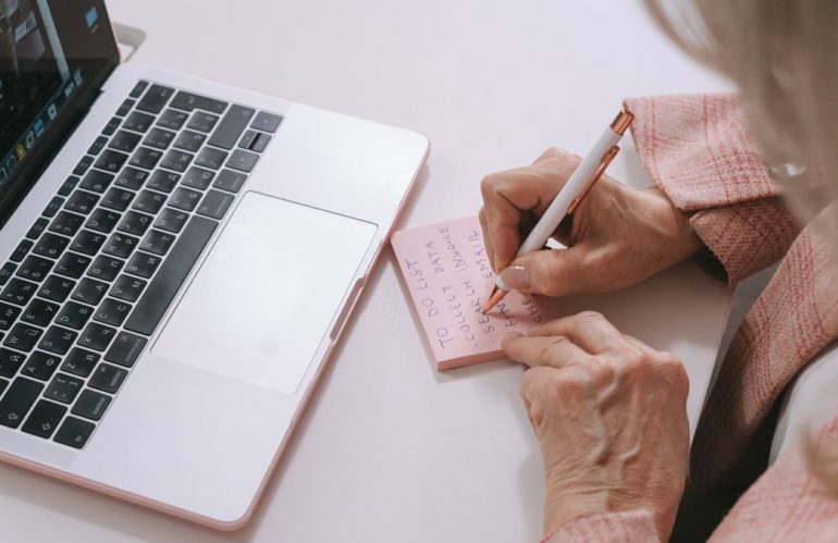Close-up of a woman writing on a pink sticky note beside a laptop. Productive work setting.
